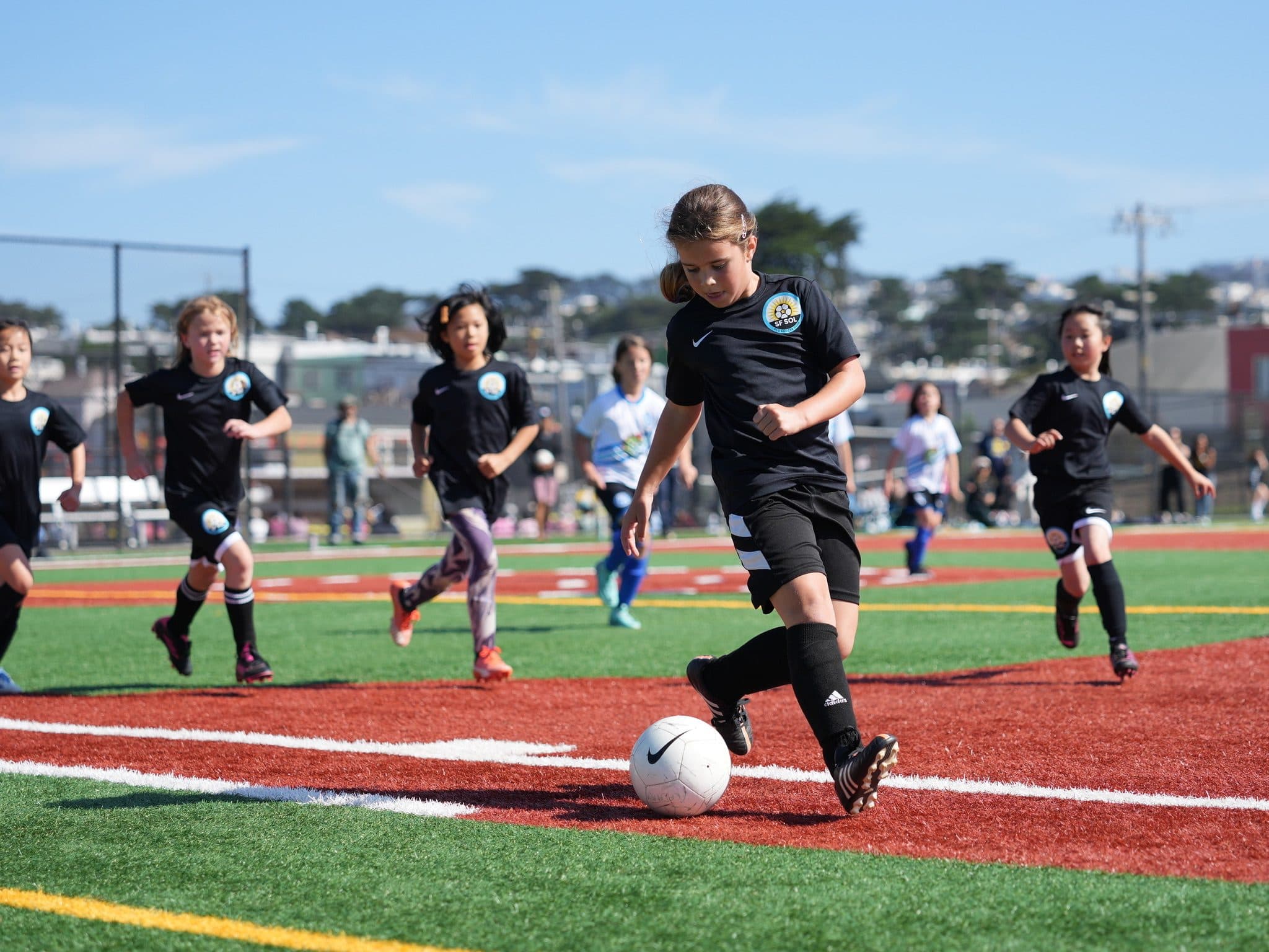 A player in a black GLG kit leads a pack of chasing players as she runs onto the ball on a sunny turf field, SF hillside houses in the background