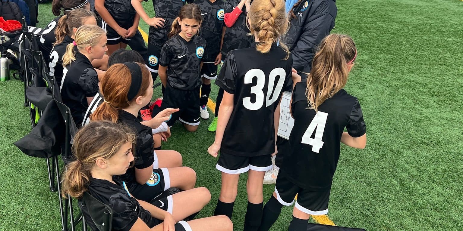 A coach in a dark jacket stands among players in black number kits during a sideline break, girls seated in camp chairs and standing in a loose huddle on an overcast game day