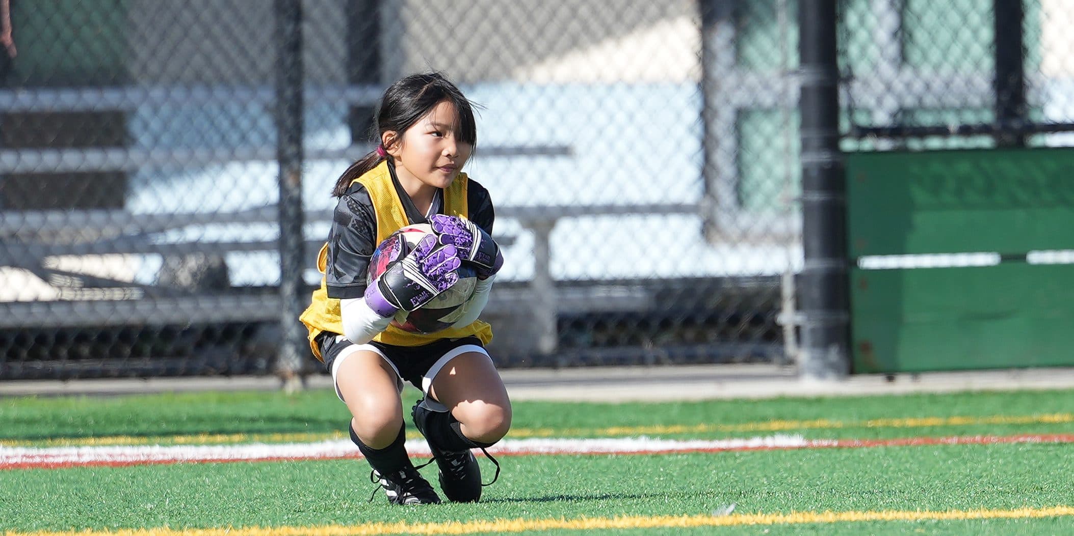 A goalkeeper in a yellow bib over a dark long-sleeve crouches to scoop up the ball, eyes locked upfield
