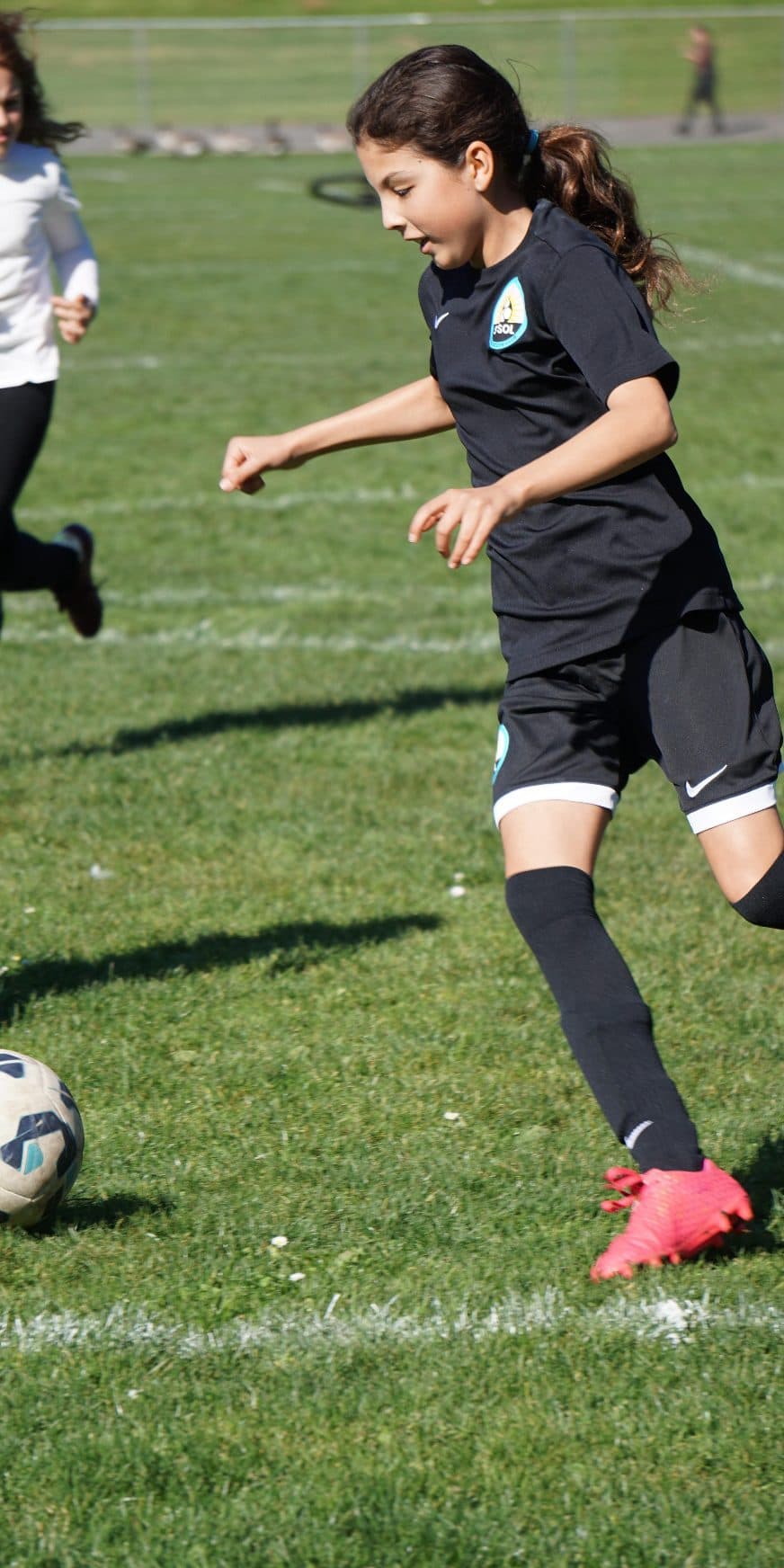 Vertical action shot of a GLG player in a black kit and red cleats winding up on the ball, hair streaming, with opponents in white trailing behind on a sunlit grass pitch